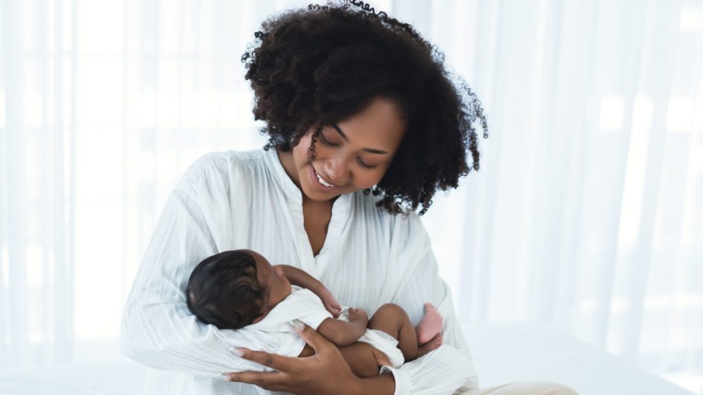 Beautiful African American young adult mother sitting on bed and holding newborn baby child in her arms, looking down at him smiling. Happy Black family lovely, nursery breastfeeding mother’s day.