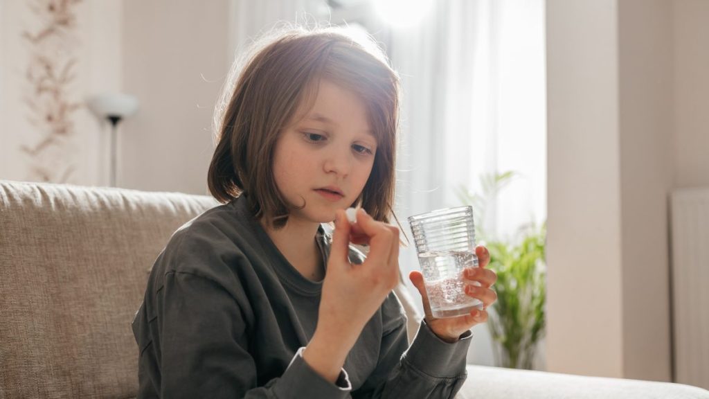A teenage girl sits on a sofa and takes a pill for illness or vitamins to maintain immunity.