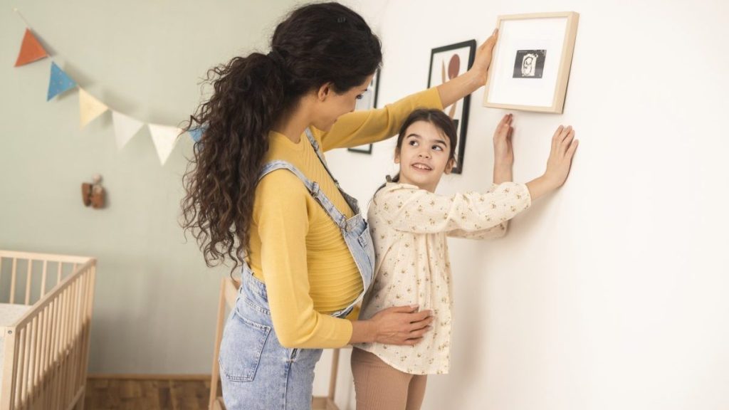 Young pregnant woman and her little daughter hanging pictures on the wall of the baby's bedroom