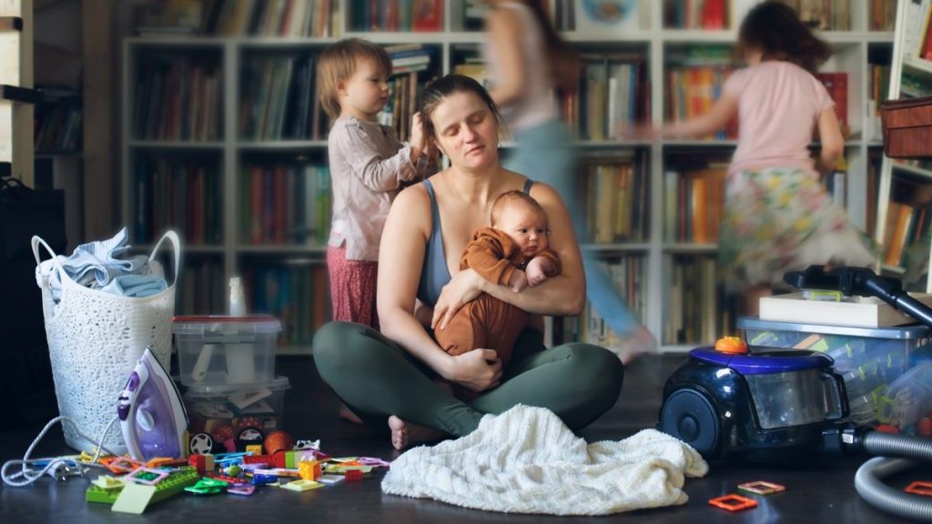 Tired, burnt-out mother with baby in her arms and out of focus children run around. Mother is sitting on floor in living room, among vacuum cleaner, laundry and children running around.