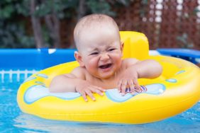 A newborn baby of 10-12 months swims in a yellow swimming ring, crying because of their fear of water. Frame swimming pool in the backyard of the house