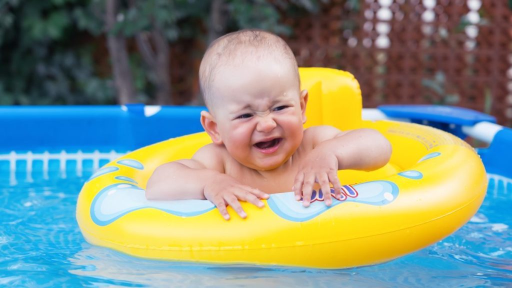 A newborn baby of 10-12 months swims in a yellow swimming ring, crying because of their fear of water. Frame swimming pool in the backyard of the house