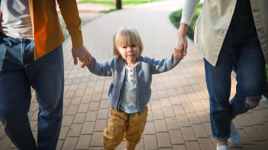 Toddler boy holds hands with both parents and swings between them on their arms during a walk in the park, showcasing how co-parenting works, which is quite different from parallel parenting.