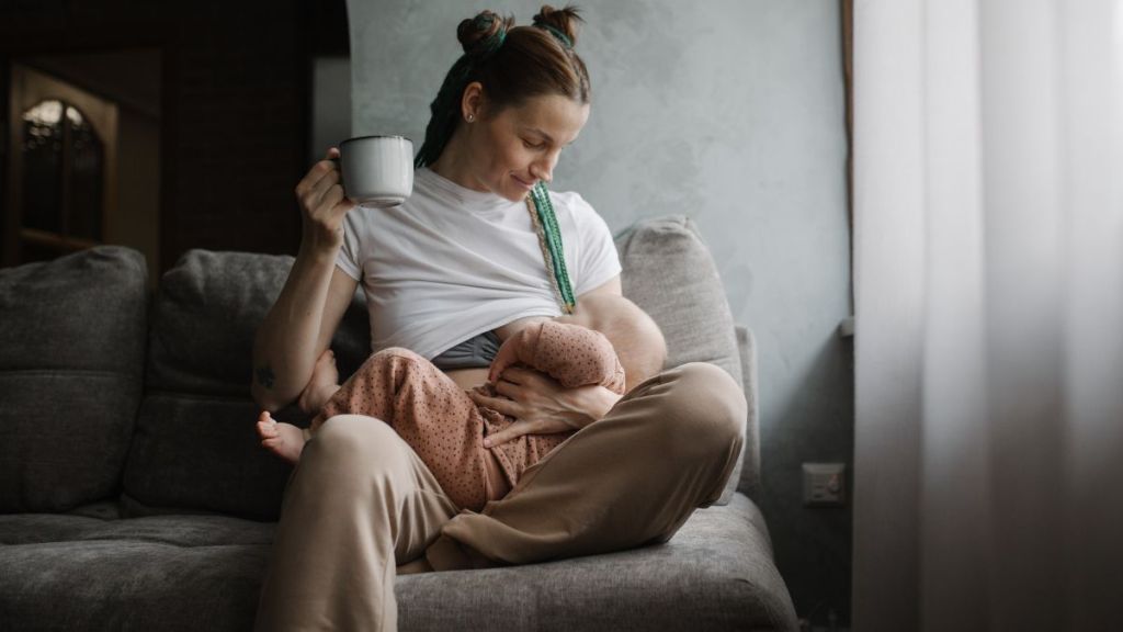 Young mother breastfeeding her baby while drinking beverage from a cup.