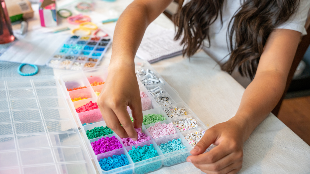 A girl sits at her kitchen table making bracelets for a fundraiser, carefully stringing beads and tying knots with a smile on her face, hoping to make a positive impact.